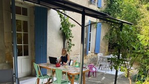 Un espacio al aire libre cubierto con mesa y sillas, una persona sentada a la mesa, y un edificio con contraventanas azules al fondo.