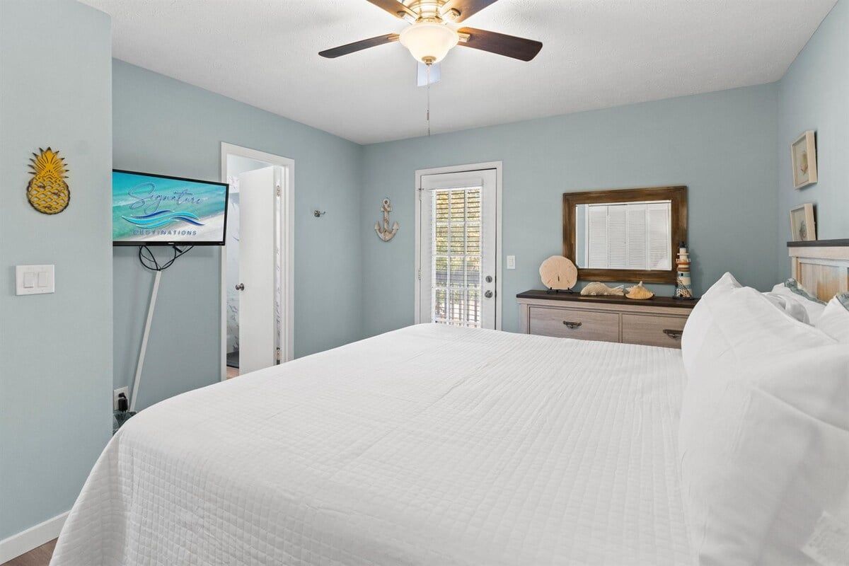 A bedroom with a bed, a ceiling fan, a television, and a door leading to another room.