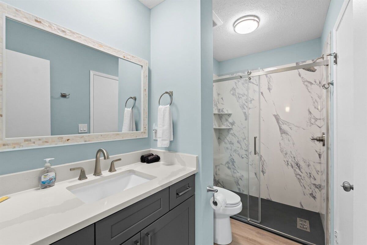 A bathroom with a white sink, a mirror, a shower with marble walls, and a toilet.