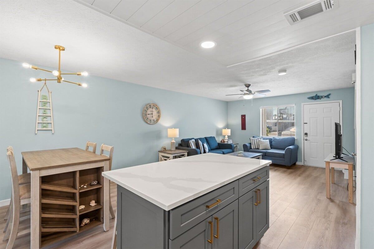 Une cuisine avec un îlot central, une table à manger avec des chaises, un canapé bleu, un téléviseur posé sur une table d’appoint et un ventilateur de plafond.