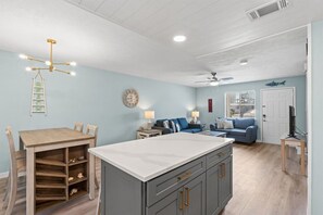 A kitchen with a central island, a dining table with chairs, a blue sofa, a TV on a side table, and a ceiling fan. - 2bd Sleeps 6 Walk to Beach Pool Newly Renovated (Panama City Beach)