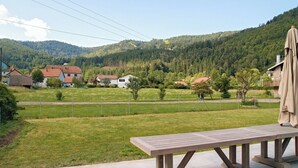 A wooden bench in a grassy area with a view of houses and a forested hill in the background.