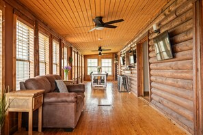 Ein Holzblockhaus-Interieur mit Holzbalkendecke, Deckenventilatoren, einem Ledersofa, einem Esstisch und einem an der Wand montierten Fernseher.