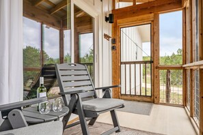 A wooden-framed room with a view of trees, a chair, a table, and a bottle of wine. - Camp Jackalope | Outdoor Trails | Fire Pit | Views (Wimberley)