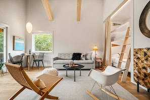 Una sala de estar moderna con sofá, mesa de centro y sillas, un comedor con mesa y sillas, y un ventanal con vistas a árboles.