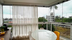 A balcony with a table and chairs, a white curtain, and a view of buildings and trees. - Aquarius Residence - Porto de Galinhas (Ipojuca)