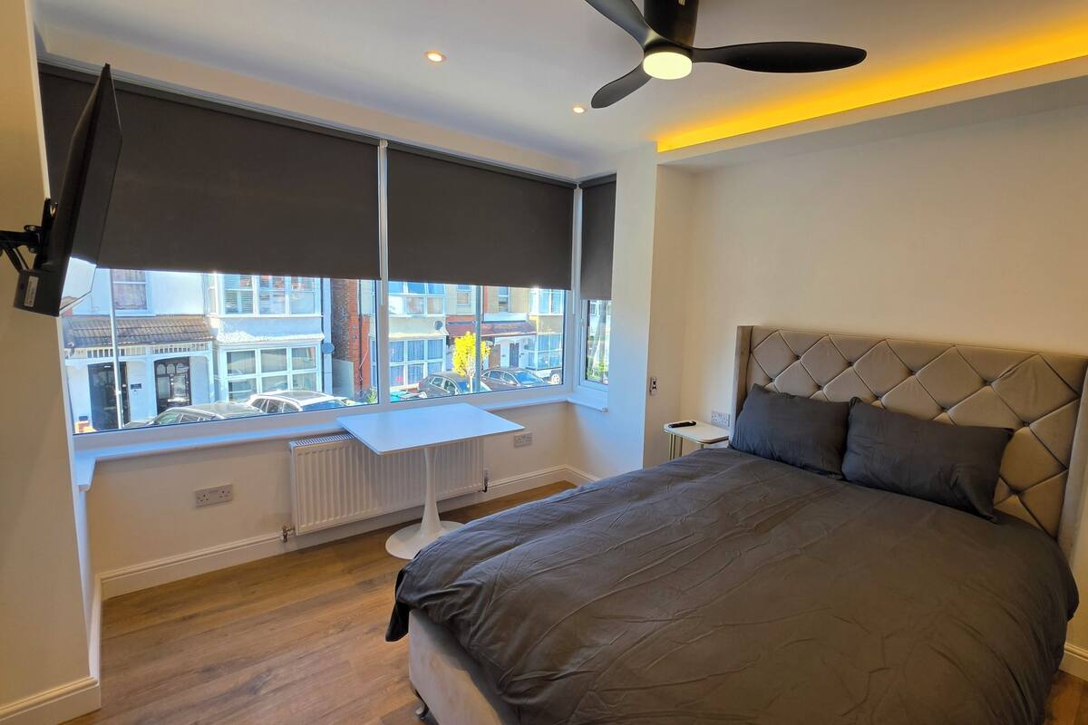 Ein Schlafzimmer mit Bett, Deckenventilator, Fenster mit Jalousien, Heizkörper und einem kleinen Tisch.