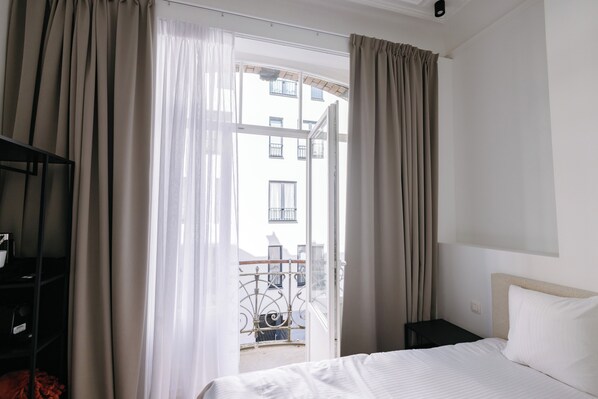 A hotel room with a bed, a nightstand, a shelf with items, and a view of a building through sheer curtains.