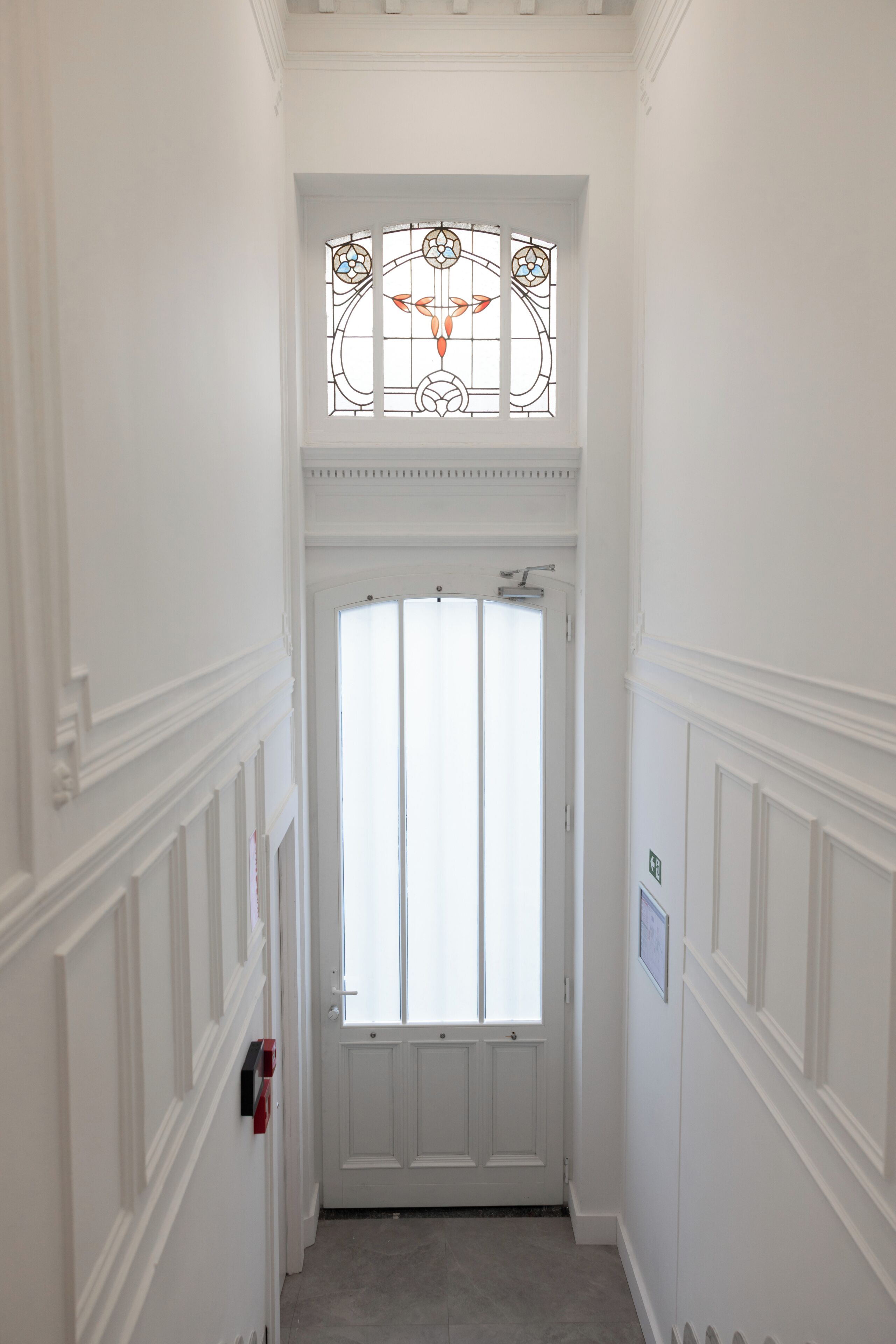 A narrow hallway with a white door, a stained glass window above, and a fire alarm on the wall.