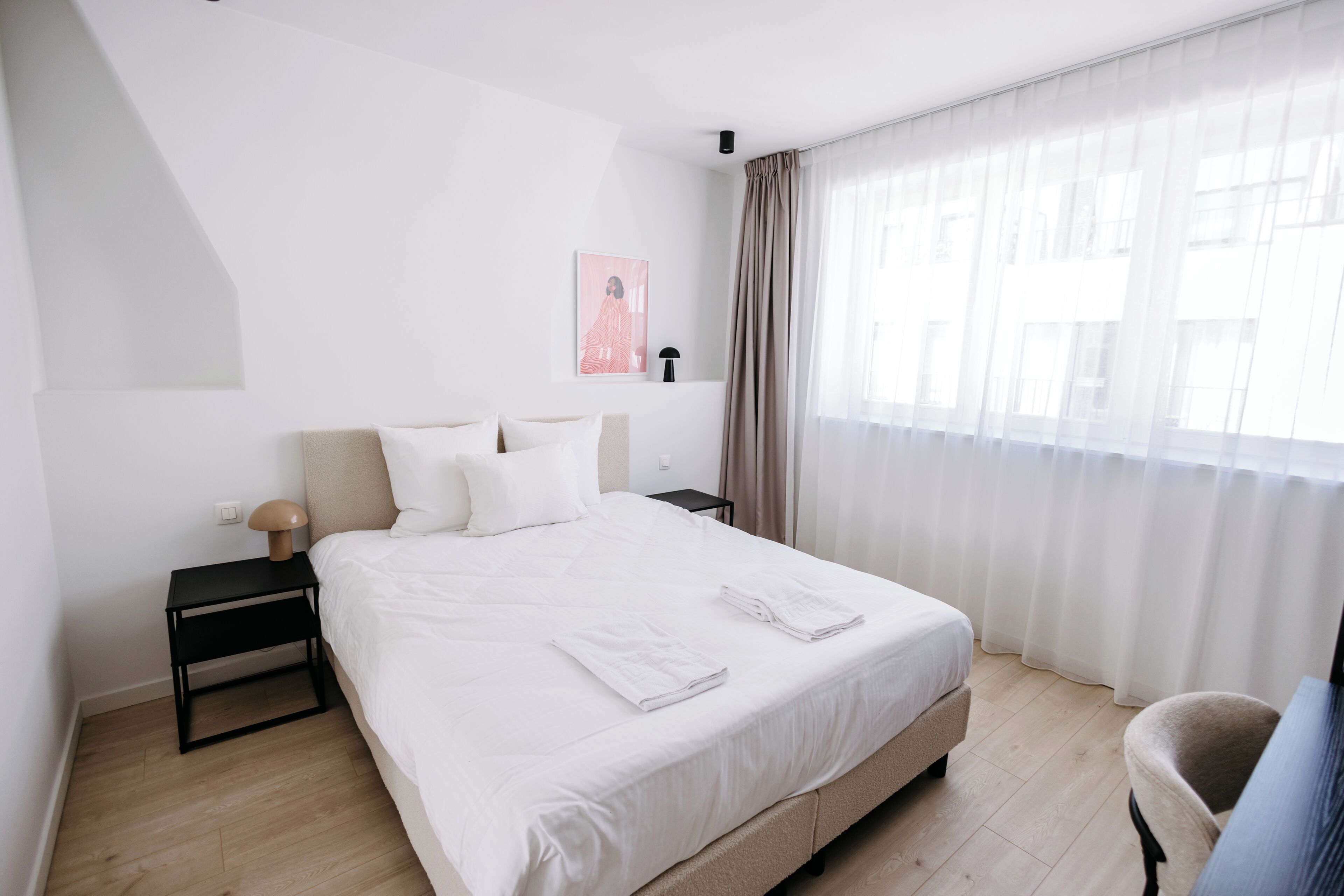 Una habitación de hotel con una cama grande, una mesita de noche, una lámpara, una silla y un ventanal con cortinas.
