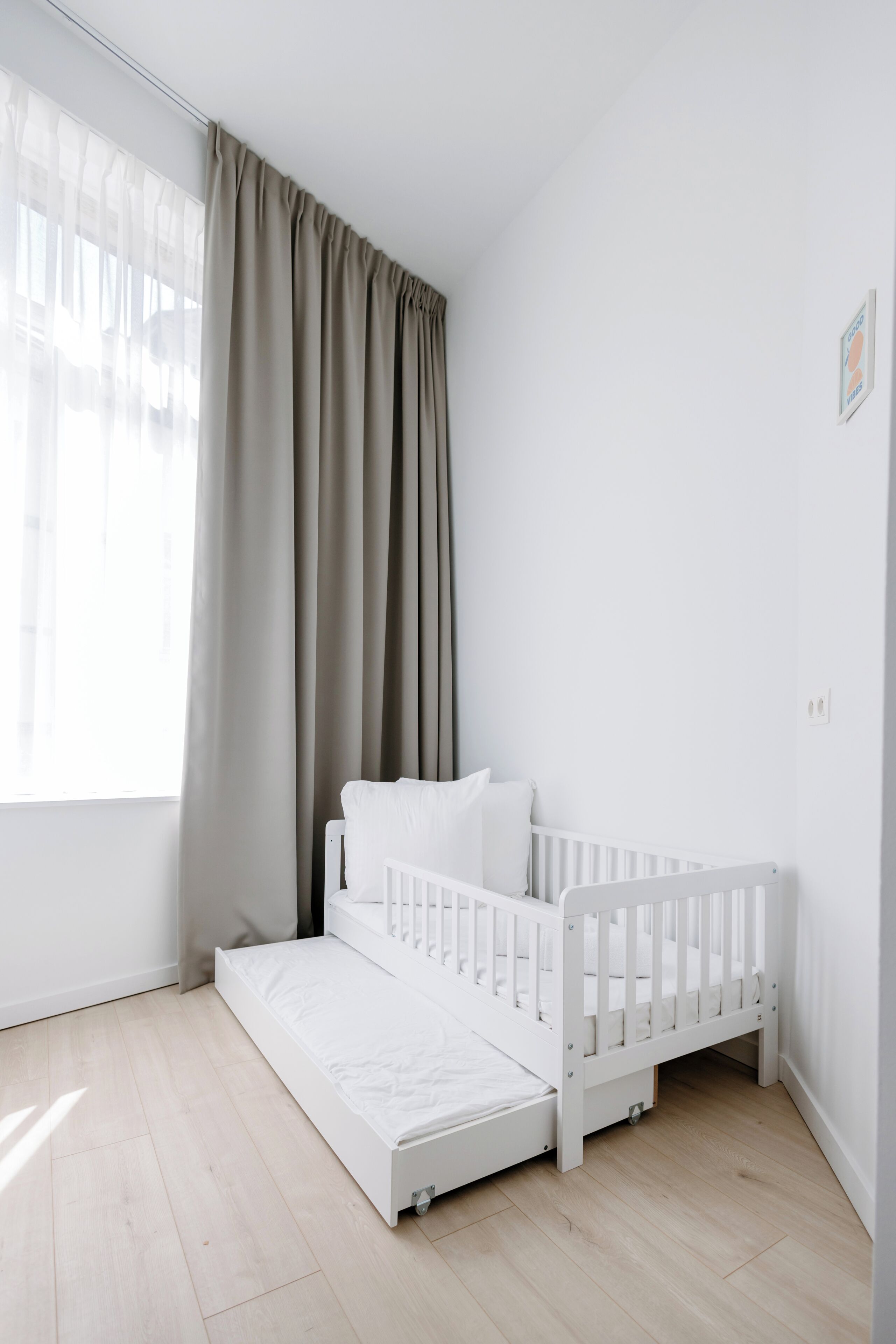 A white crib with a mattress and a small step-up platform, positioned against a white wall with a window and curtains.