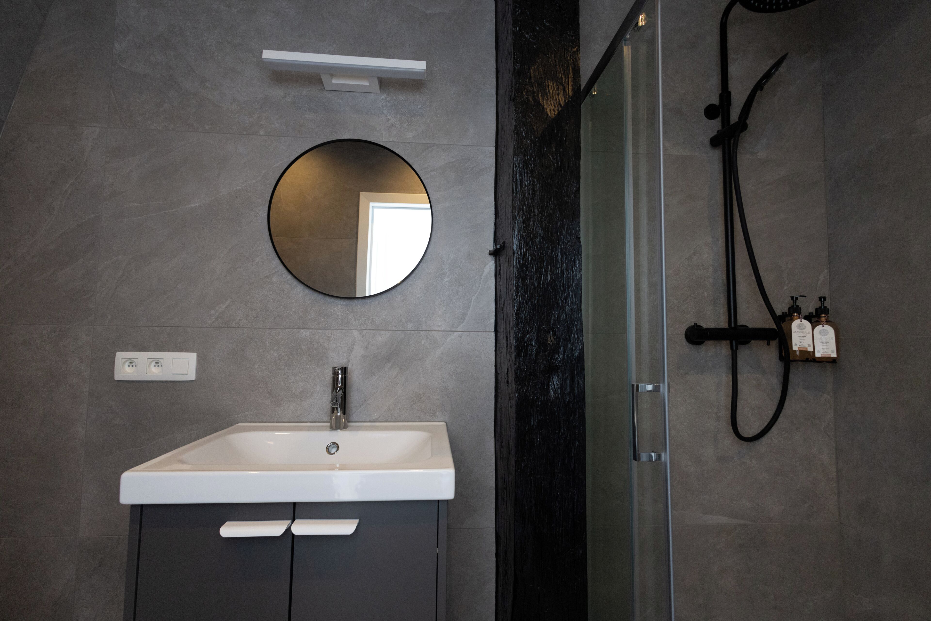 A modern bathroom with a white sink, a round mirror, a glass-enclosed shower, and a wall-mounted soap dispenser.