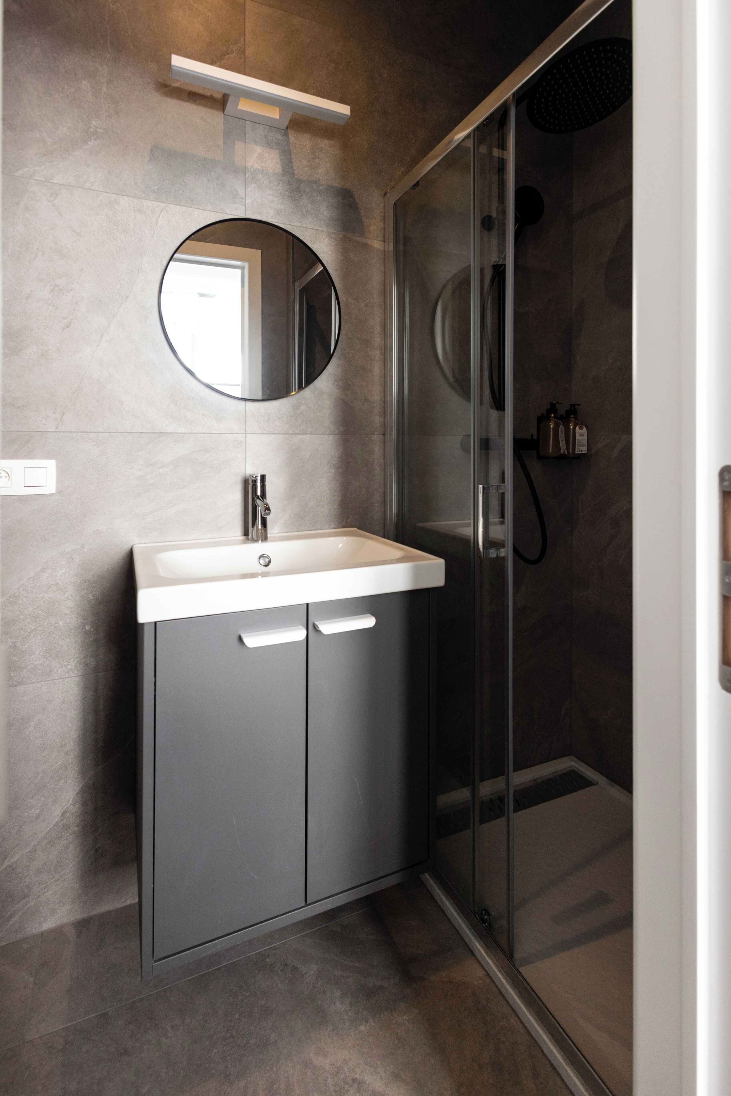 Un baño moderno con ducha envuelta en vidrio, un espejo redondo sobre el lavamanos y un mueble gris con dos cajones.
