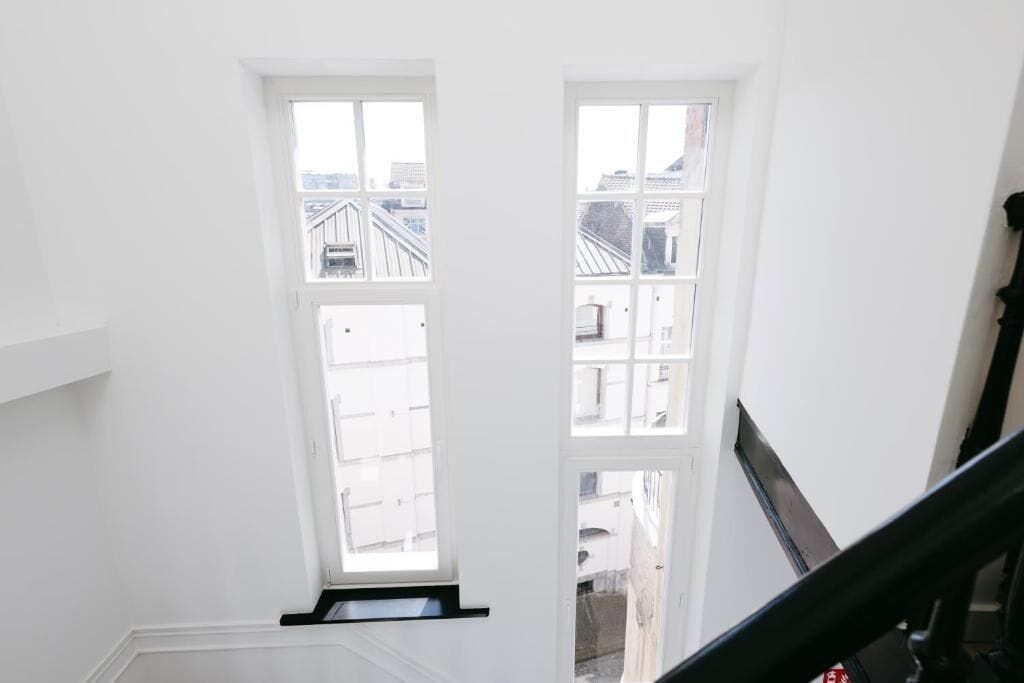 A high-ceiling room with two large windows, a black staircase, and a white wall.