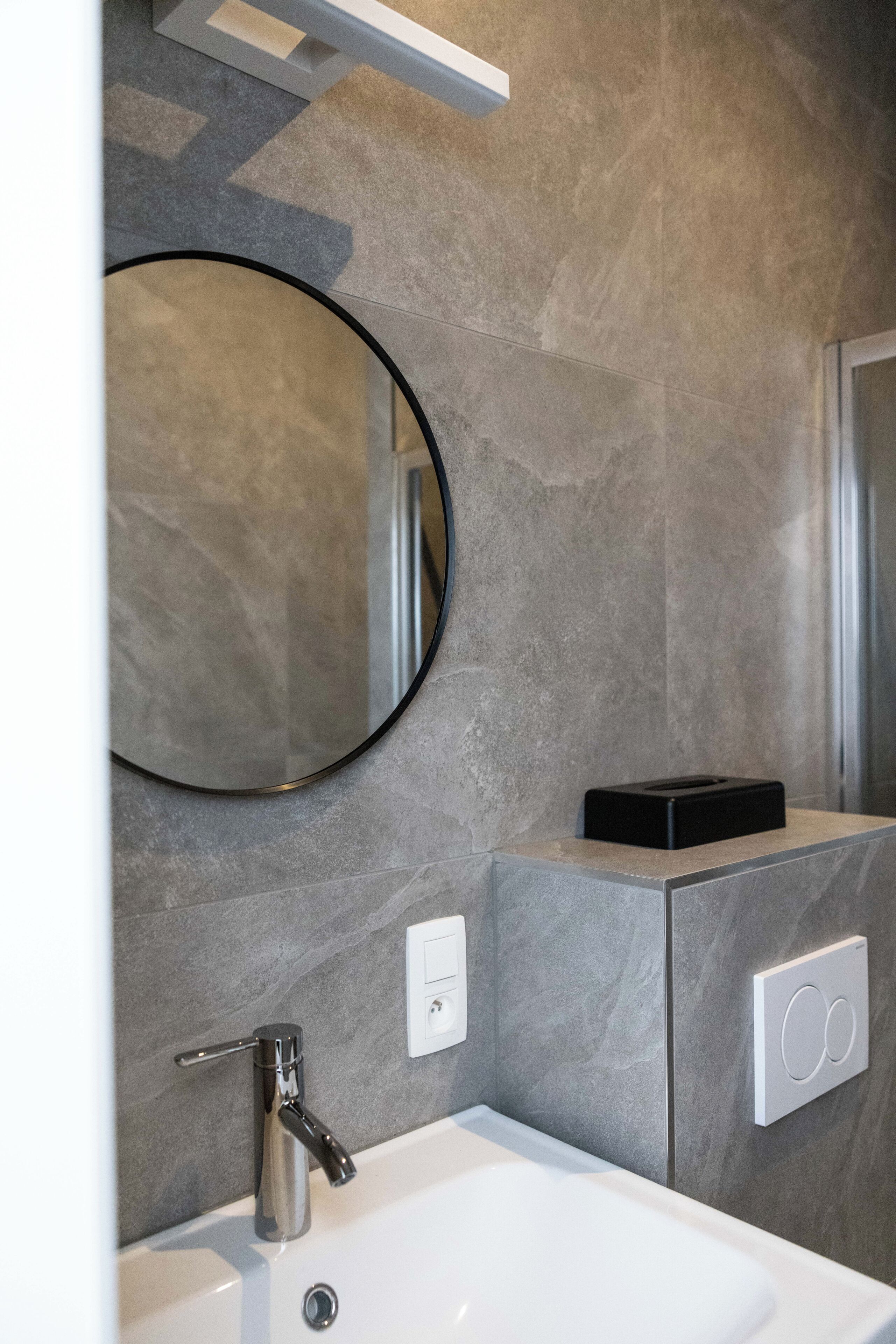 A bathroom with a white sink, a round mirror, and a black soap dish on a grey tiled wall.