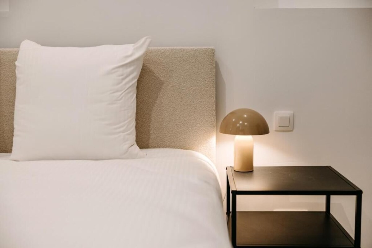 A bed with a white pillow and a beige headboard, a bedside table with a lamp, and a power outlet on the wall.
