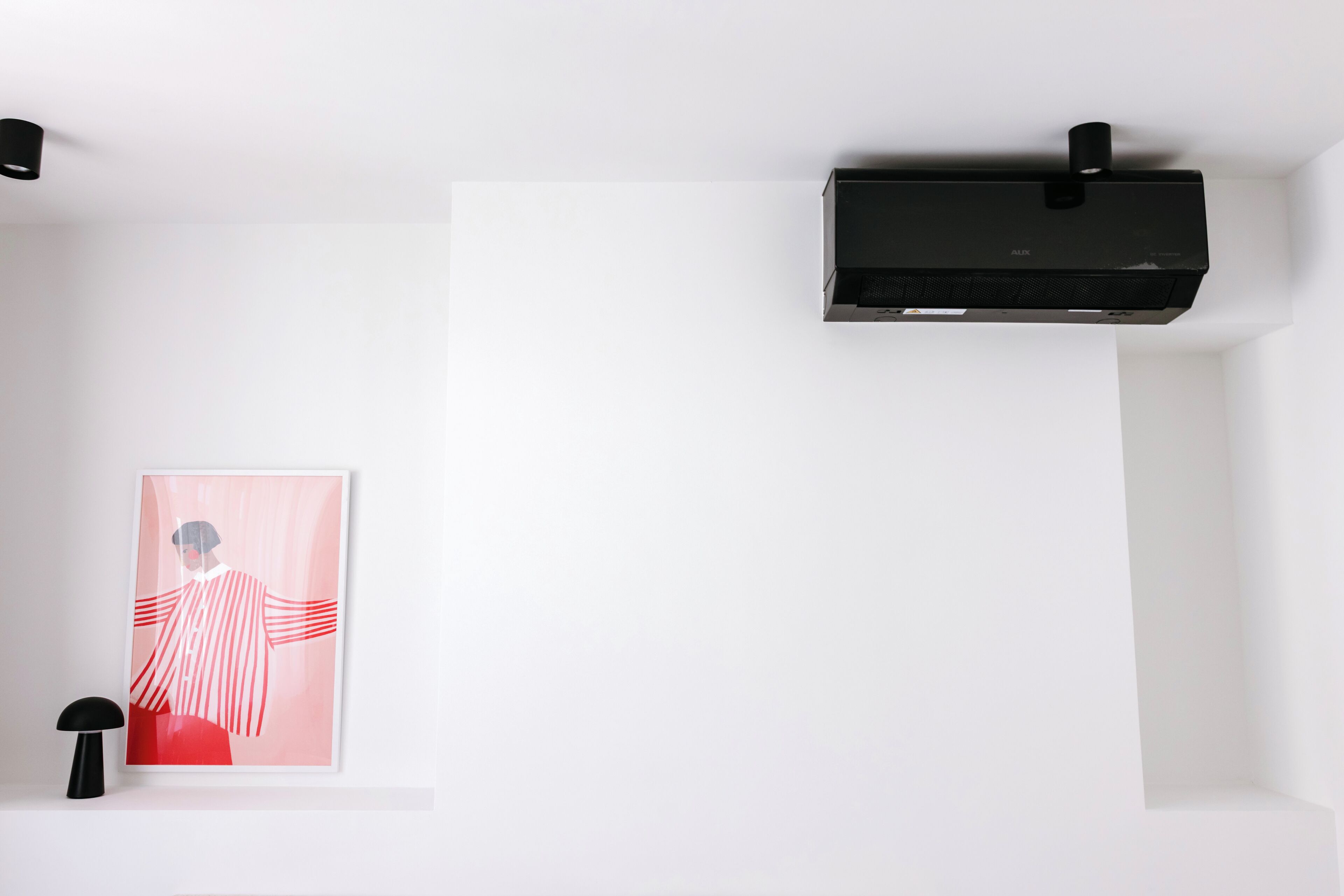 A white room with a black air conditioning unit, a framed abstract painting, and a black floor lamp.