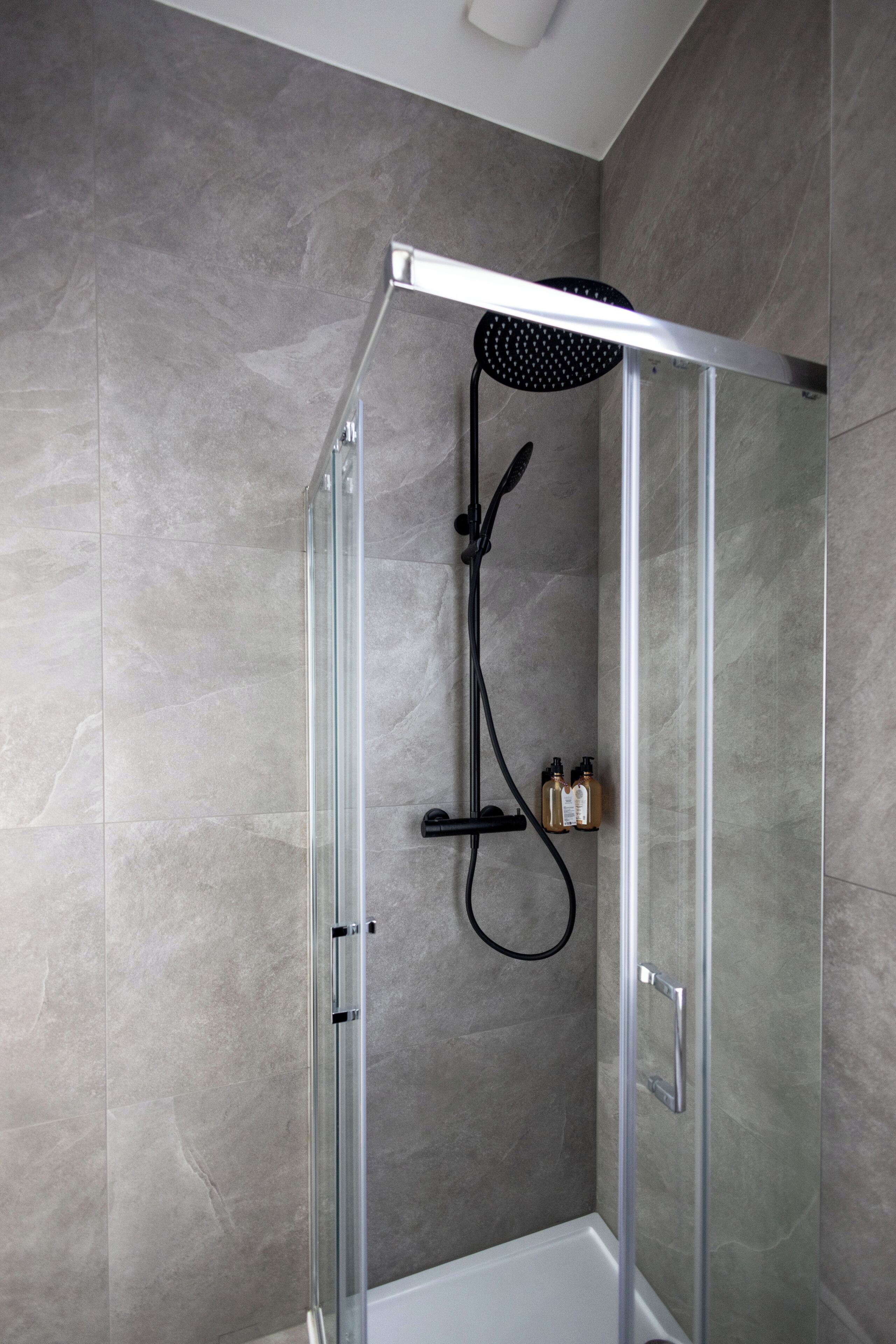 A glass-enclosed shower area with a handheld showerhead, a wall-mounted faucet, and two bottles of shampoo.