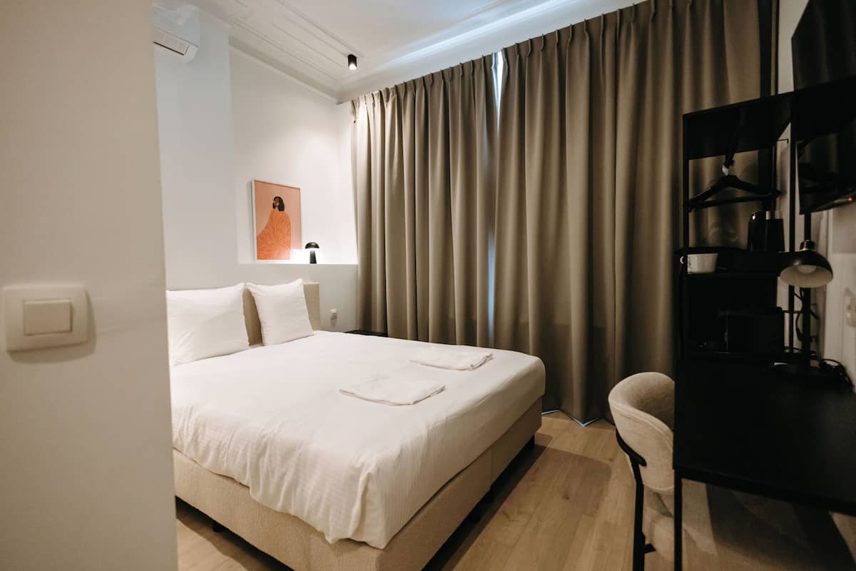 Une chambre d’hôtel avec un lit, un bureau, une chaise, une lampe et une fenêtre avec des rideaux.