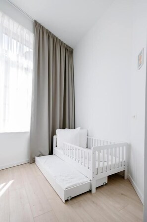A white crib with a mattress and pillows, a window with sheer curtains, and a light wooden floor.