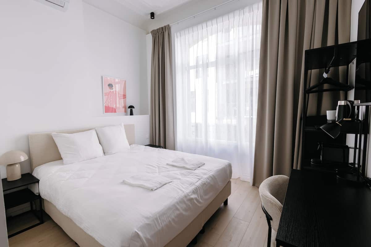 Une chambre d’hôtel équipée d’un lit, d’un bureau, d’une chaise, d’un porte-manteau et d’une fenêtre avec des rideaux.