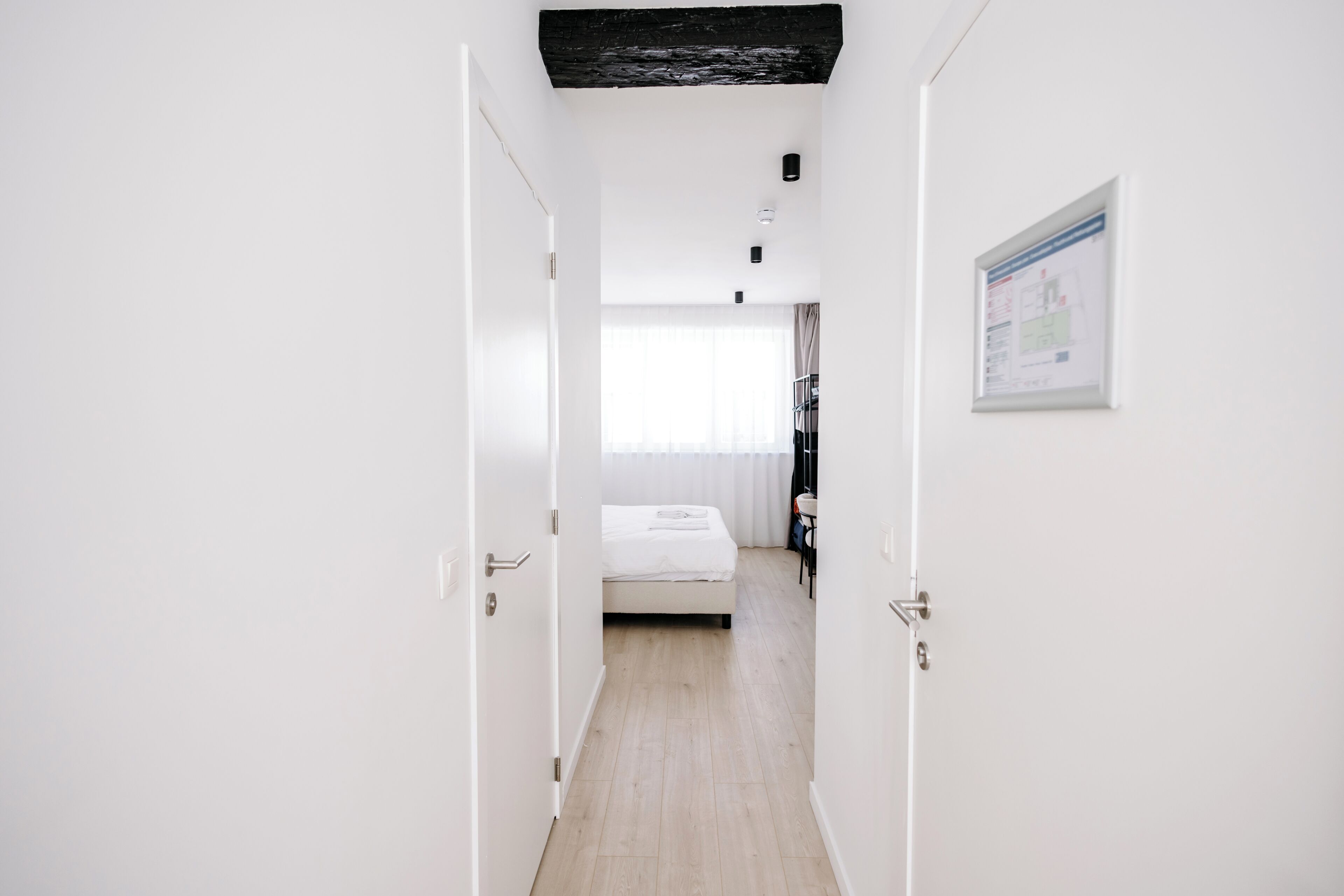 A narrow hallway leading to a bedroom with a bed, a window with sheer curtains, and a framed chart on the wall.