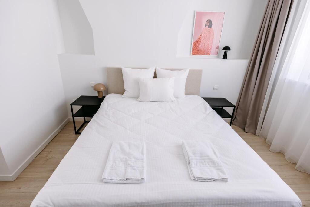 A neatly made bed with white linens, two bedside tables with lamps, a framed picture on the wall, and a window with curtains.