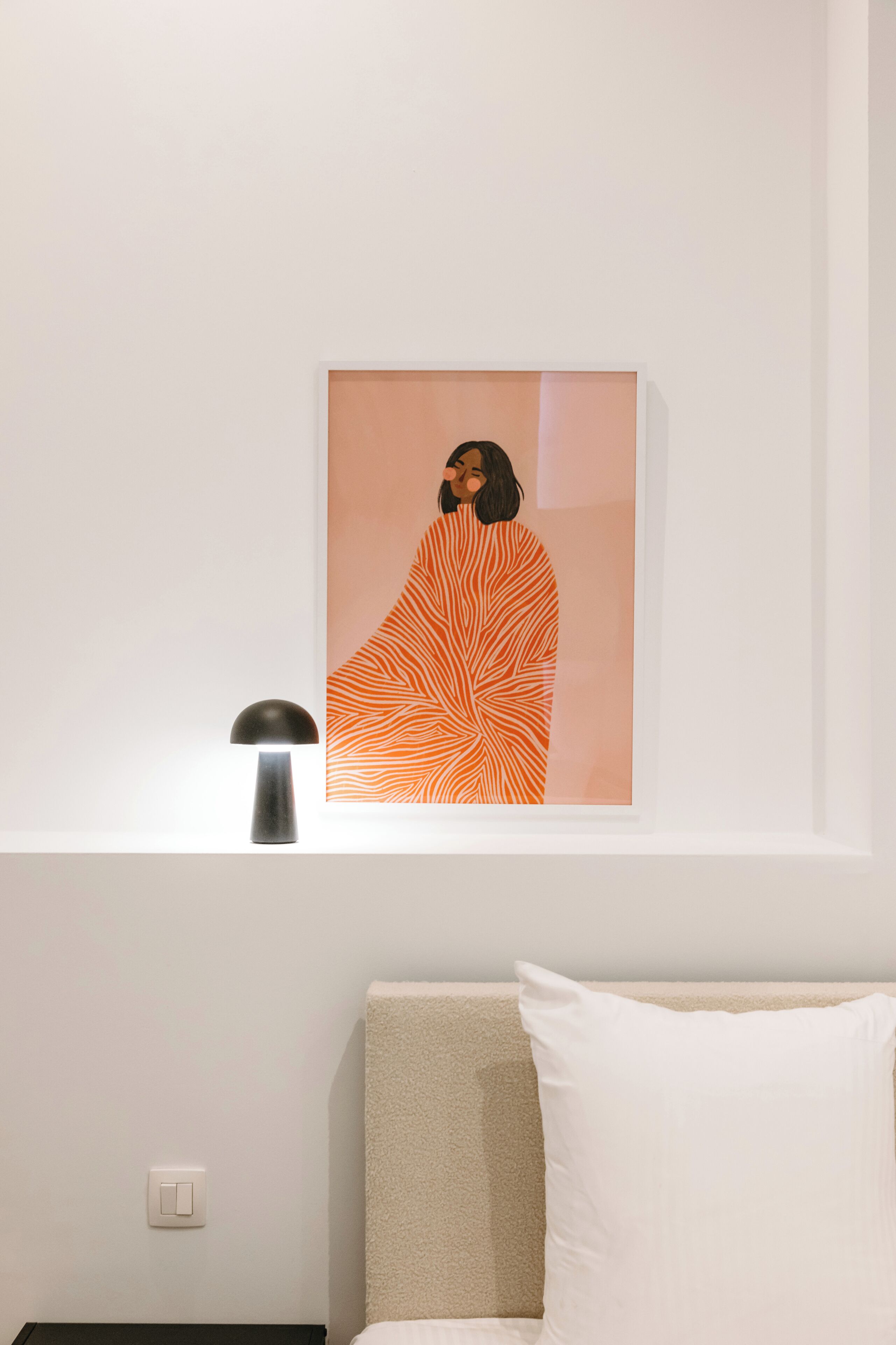 Una obra de arte en un marco en la pared, una lámpara de noche y una cama con un almohadón.