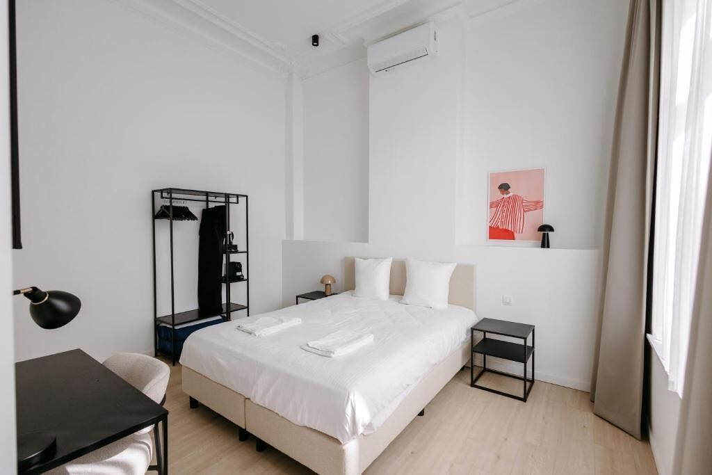 Quarto de hotel com cama, escrivaninha, cadeira, mesinha, guarda-roupa e um quadro na parede.