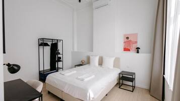 Quarto de hotel com cama, escrivaninha, cadeira, mesinha, guarda-roupa e um quadro na parede.