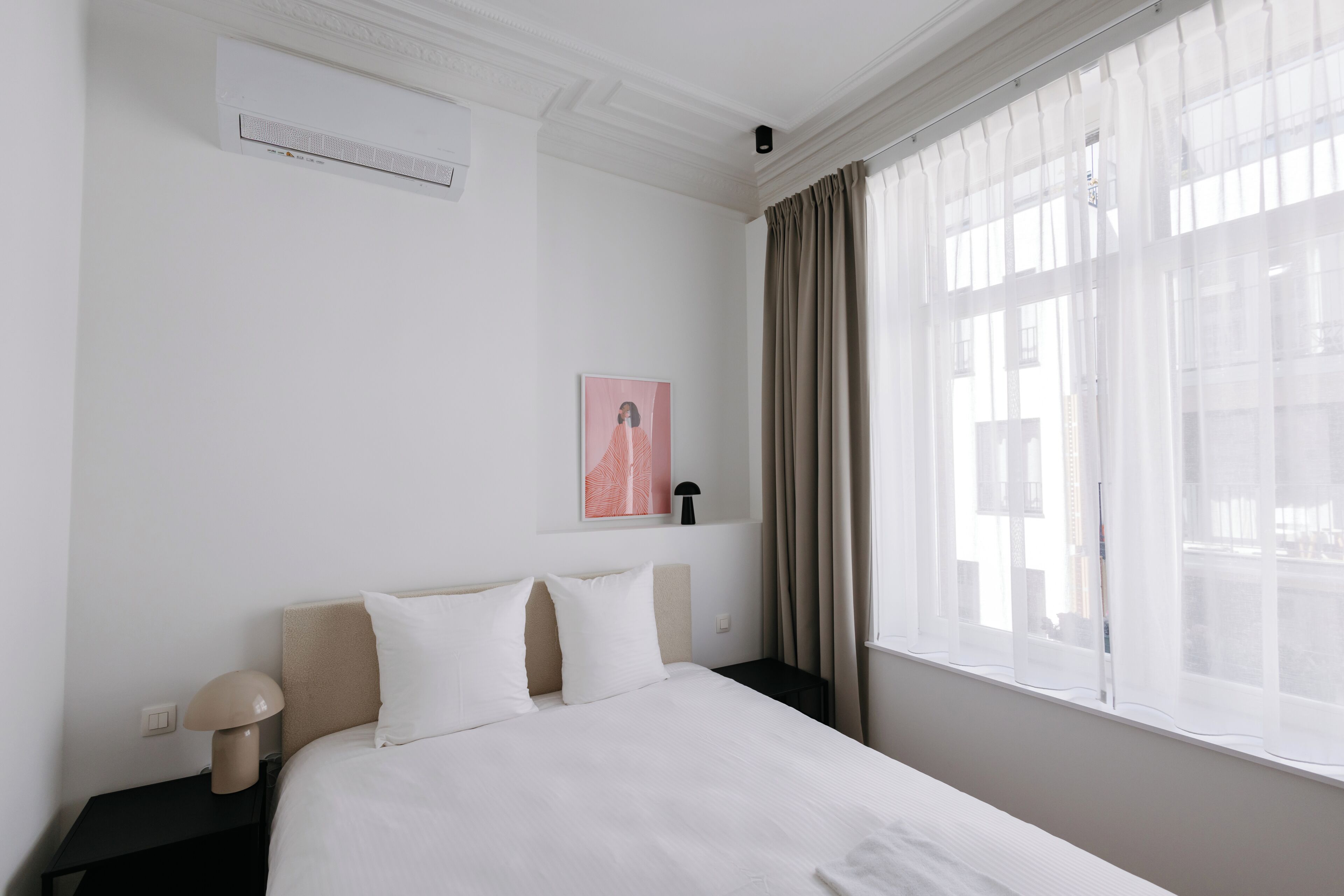 Ein Hotelzimmer mit Bett, Nachttisch, Lampe, einem Bild an der Wand und einem Fenster mit Vorhängen.