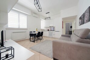 Ein modernes Wohnzimmer mit einer Couch, einem Esstisch mit Stühlen, einer Küchenzeile und einem großen Fenster.