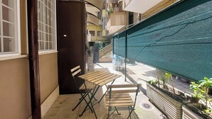 Un estrecho pasillo exterior con una mesa de madera, sillas, un armario negro y una pantalla verde.