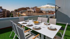 Eine Dachterrasse mit Esstisch, gedeckt für vier Personen, umgeben von Stühlen, einem großen Sonnenschirm und Blick auf die Stadtlandschaft.