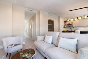 Ein modernes Wohnzimmer mit einer Couch, einem Sessel und einem Couchtisch. Es gibt eine Küchenzeile mit einer Hängeleuchte.