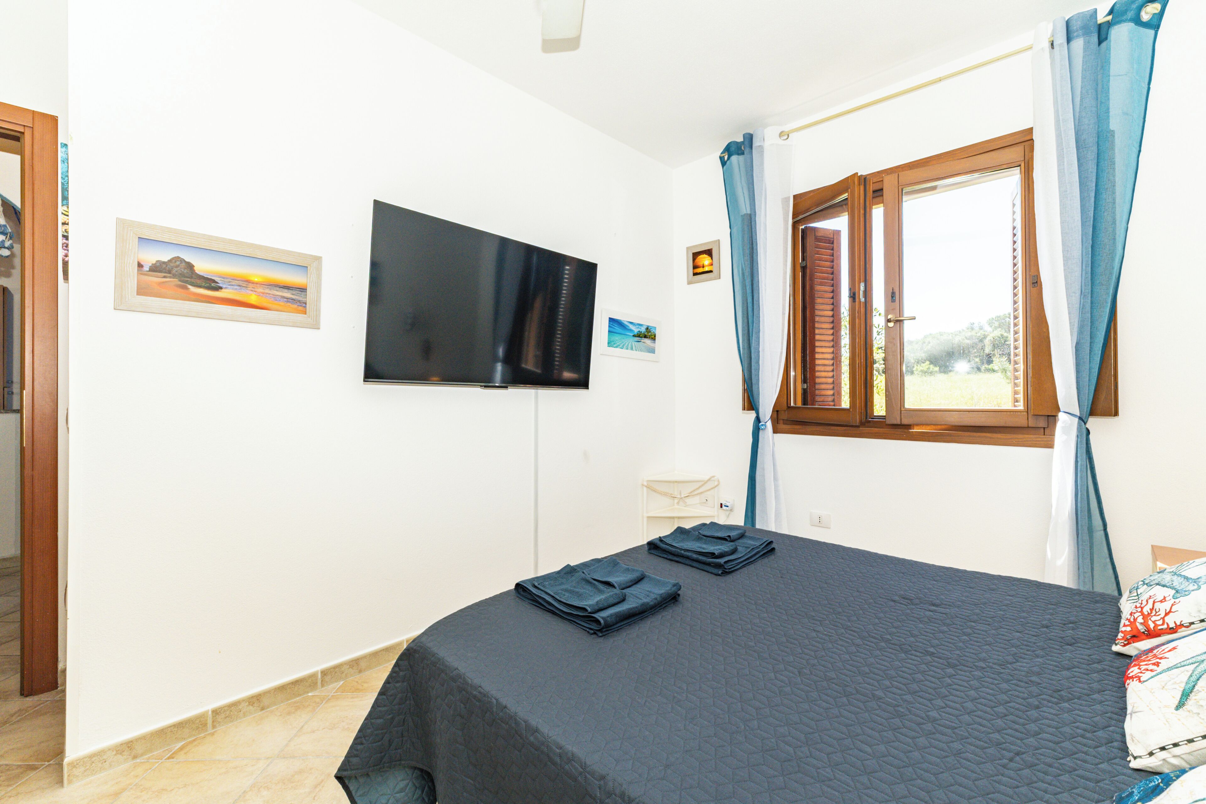 Ein Schlafzimmer mit Bett, Wandfernseher, Fenster mit Vorhängen und einem kleinen Tisch.