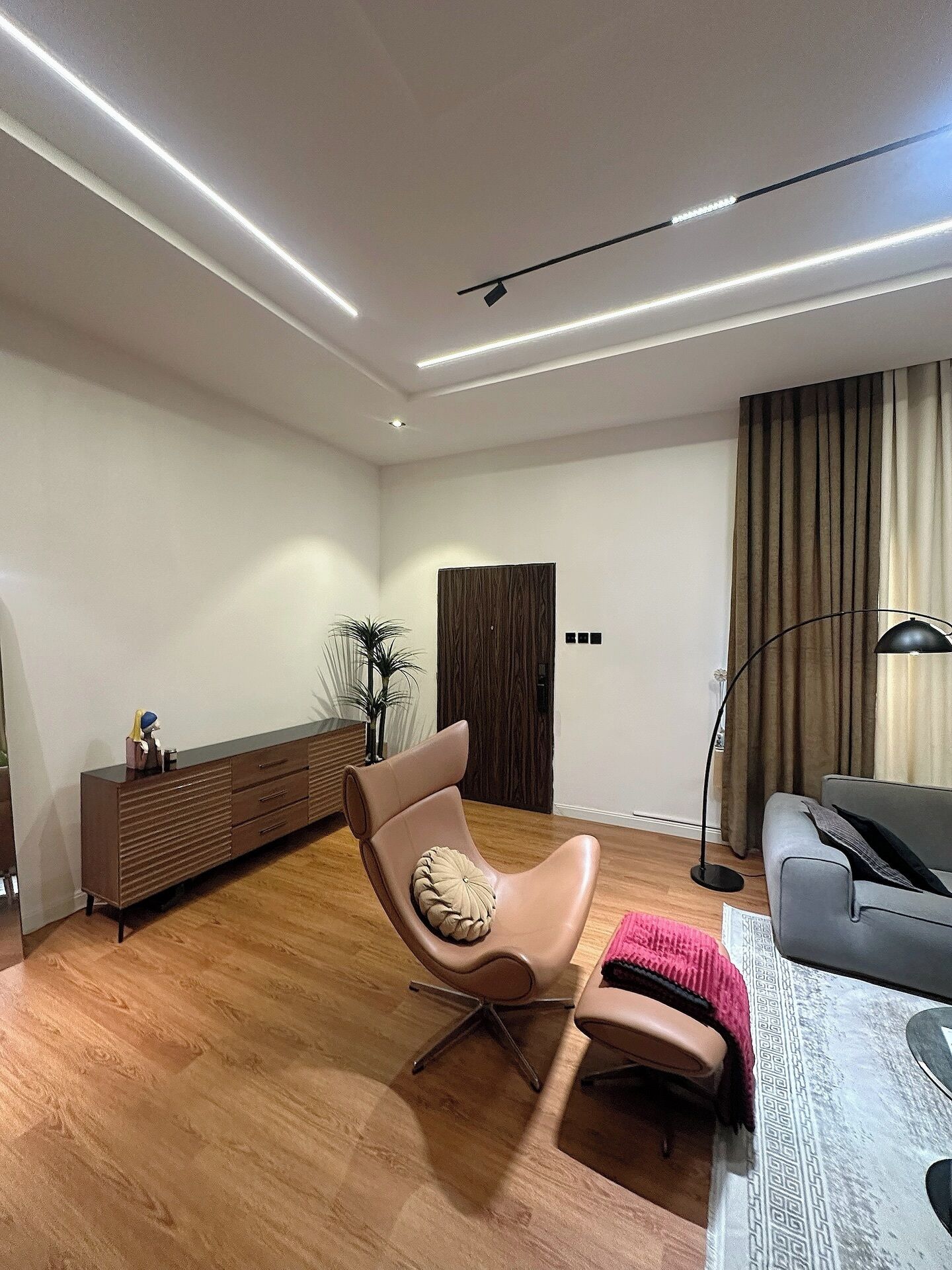 A modern living room with a leather armchair, a grey sofa, a wooden floor, a dark brown cabinet, and a floor lamp.