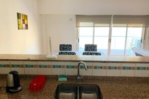 Eine Küche mit zwei Spülen, einem Wasserkocher, einer roten Seifenschale und gemusterten Stühlen nahe einer Glastür.