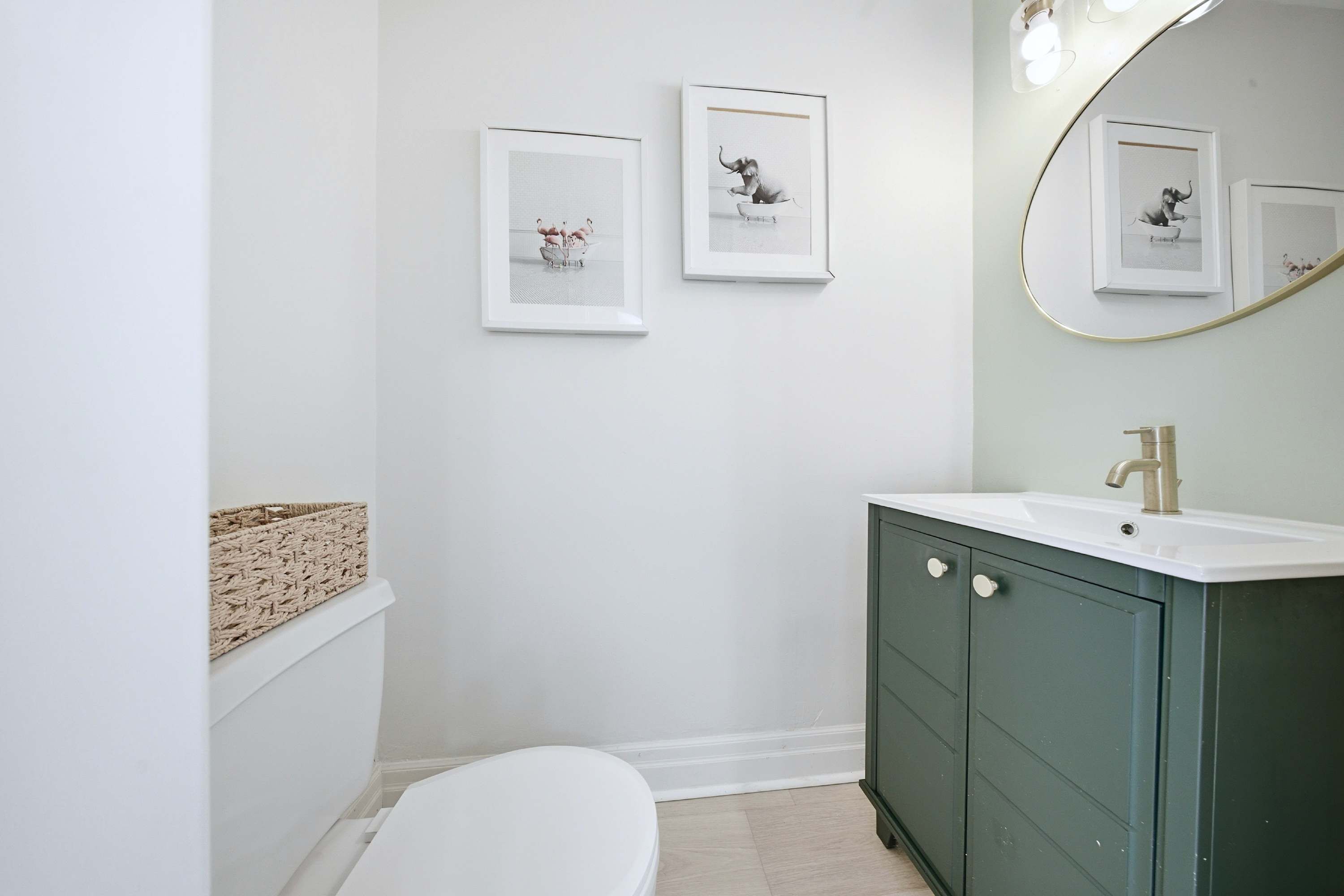 Une salle de bain avec des toilettes blanches, un meuble vert, un lavabo blanc et deux tableaux encadrés accrochés au mur.