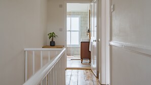 Un pasillo estrecho con suelo de madera, barandilla blanca, planta en maceta y una puerta que da a un baño con pared de azulejos y ventana.