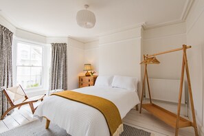 A bedroom with a bed, a wooden swing, a window with curtains, a nightstand with a lamp, and a radiator. - The Tide House, Broadstairs (Broadstairs)