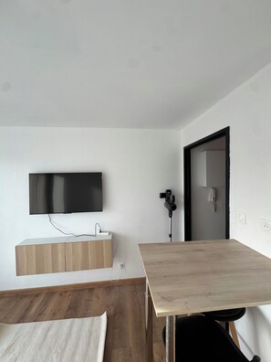 A modern room with a flat-screen TV mounted on the wall, a wooden desk, a white bedside table, and a black chair. - L'Escapade Alsacienne near Europa Park (Erstein)