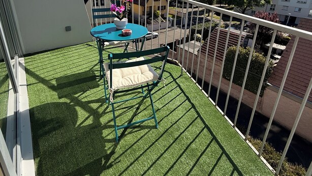 Een balkon met kunstgras, een tafel met stoelen, een bloemenvaas en uitzicht op omliggende gebouwen.