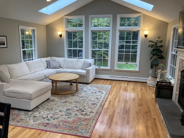 A loft-style living room with a sectional sofa, a round coffee table, a patterned rug, and large windows with a view of greenery.