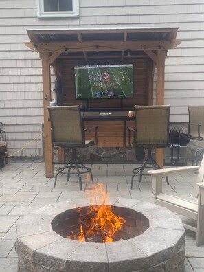 A patio with a stone fire pit, outdoor seating, and a TV mounted on a wooden pergola. - World Cup Ready Home, 10 minutes to Stadium! (Foxborough)