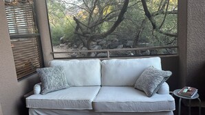Un sofá blanco con almohadones estampados, una mesita redonda con un libro y una botella, y una ventana con vista a árboles y rocas.