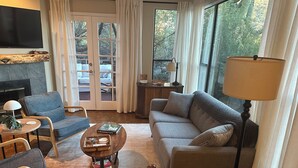 Una sala de estar con sofá, sillones, una mesa de centro, chimenea y ventanas grandes con cortinas.
