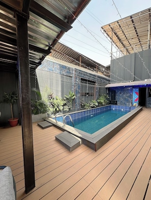 Ein Schwimmbecken mit einer gefliesten Kante, umgeben von einem Holzterrassenbelag, unter einer Metall-Dachkonstruktion.