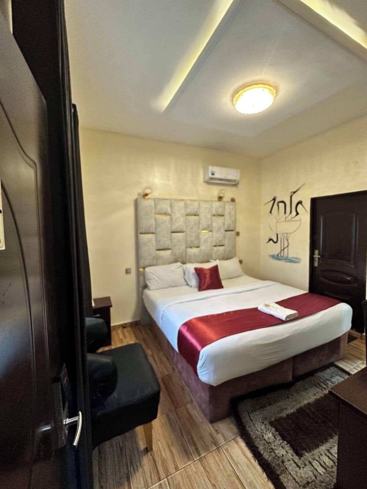 A hotel room with a bed, a bedside table, a chair, a wall-mounted air conditioner, and a door.