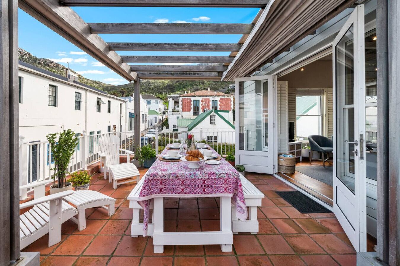 A covered outdoor dining area with a table set for two, white chairs, and potted plants.
